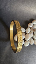 Gold bracelet with textured pattern on a dark surface with pebbles
