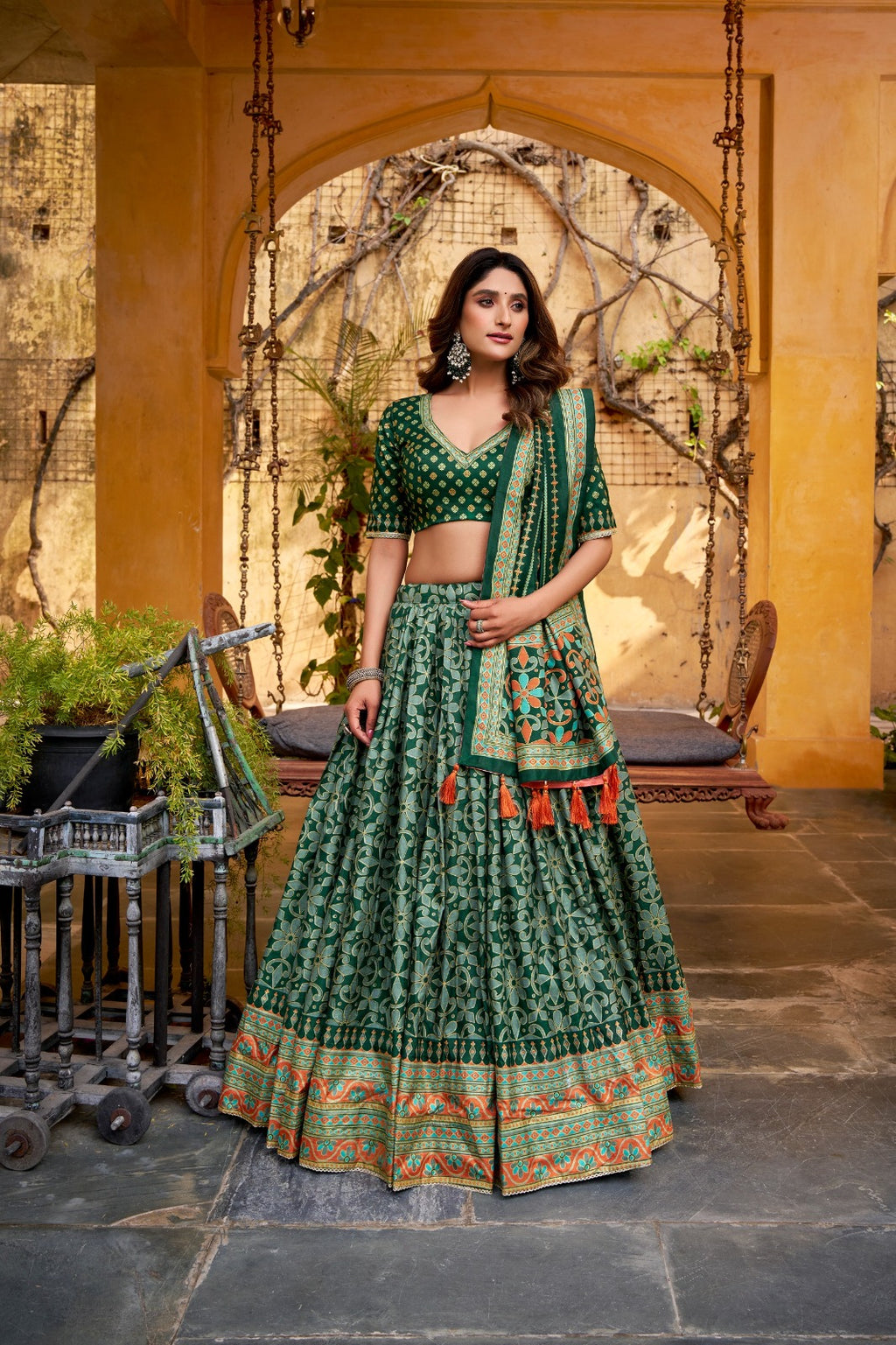 Woman in a green traditional outfit standing in an outdoor setting with a decorative arch and plants.
