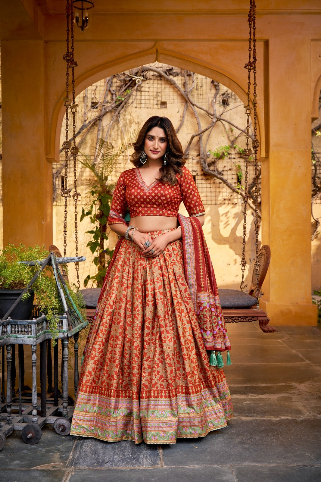 Woman in a traditional red and orange lehenga standing in an outdoor setting with arches and plants.