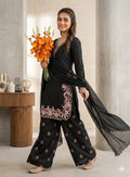 Woman in a black traditional outfit with floral patterns holding orange flowers indoors.