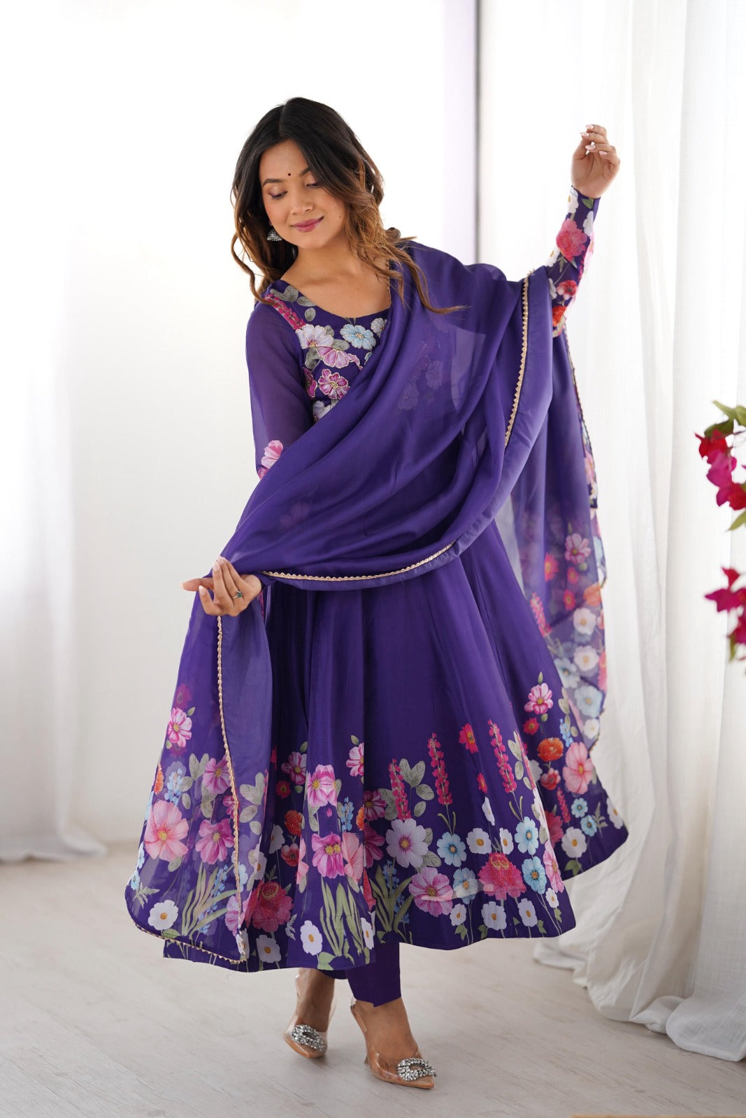 Woman wearing a purple floral dress with a matching dupatta against a white curtain background